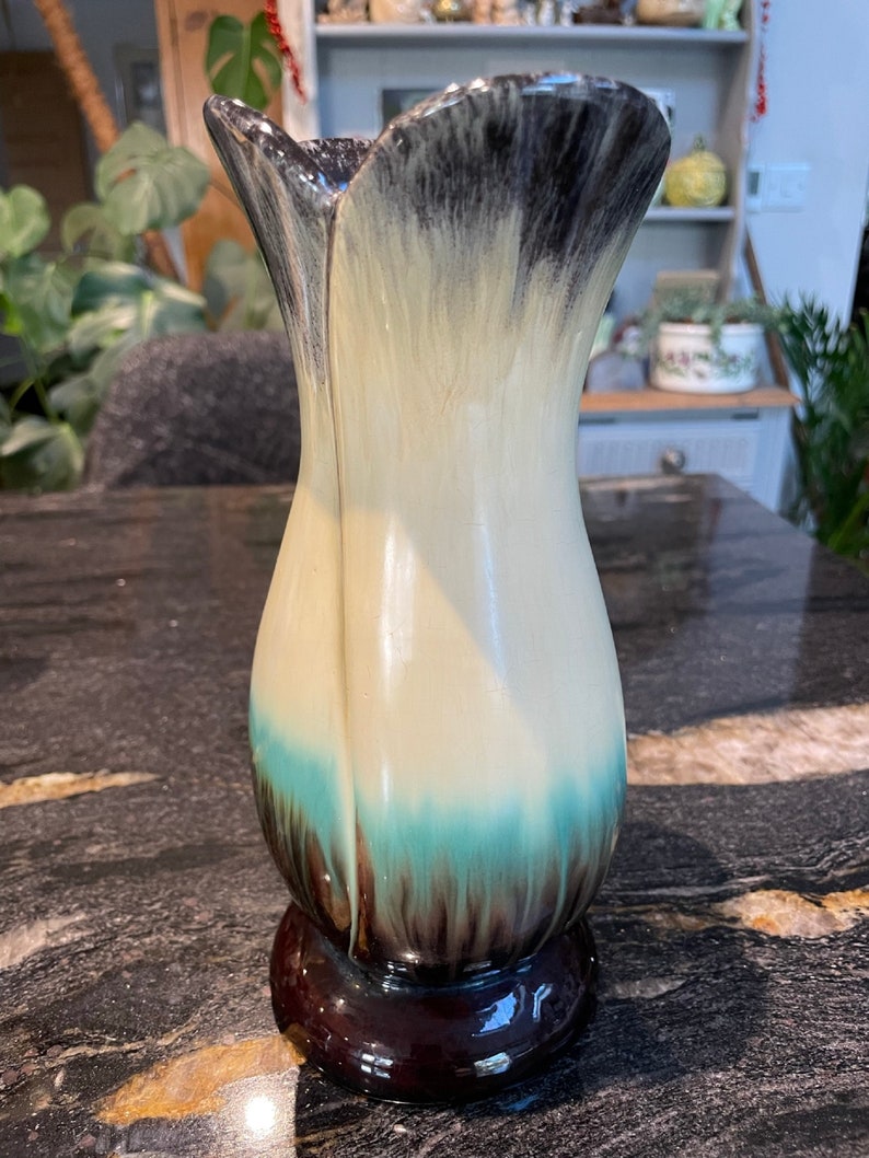 May include: A ceramic vase with a unique design. The vase is white with black and brown accents and a teal blue band around the middle. The vase has a wide mouth and a narrow base.