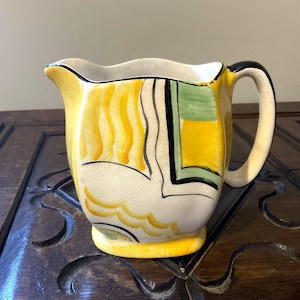 May include: A vintage ceramic jug with a yellow, cream, and black design. The jug features a curved handle and a spout. The design includes yellow wavy lines, green and black geometric shapes, and a cream-coloured base.