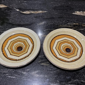 May include: Two beige ceramic plates with a concentric geometric design in shades of brown, orange, and cream. The plates have a textured rim and a dark brown centre. The plates are likely for serving food.