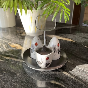 May include: A set of three white ceramic salt and pepper shakers with a floral design. The shakers are on a silver stand with a round mirror.