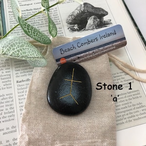 May include: A black stone with a gold symbol etched on it. The stone is sitting on a beige fabric bag with a white label that says "Beach Combers Ireland" and a small Etsy logo. The label is next to a book with text about the history of Ireland.