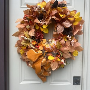 Puede incluir: Una corona de otoño circular con una mezcla de hojas artificiales naranjas, amarillas y marrones, piñas de color marrón oscuro, pequeñas bayas blancas y una calabaza decorativa central. Un gran lazo de terciopelo dorado texturizado cuelga abajo.