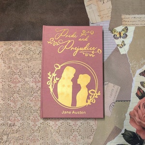 May include: A pink hardback book with a gold silhouette of two people and floral designs. The title of the book is "Pride and Prejudice" by Jane Austen.