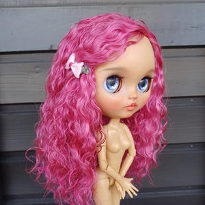 May include: A nude-colored fashion doll with large blue eyes and long, curly, bright pink hair. A small pink bow adorns the doll's hair. The doll is posed with arms bent at the elbows and hands near the torso.