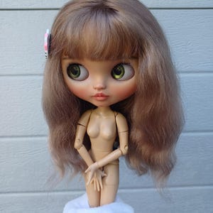 May include: A doll with shoulder-length light brown hair, a fringe, and large green eyes. The doll has a pale skin tone, a small nose, and peach-coloured lips. A small pink and white circular hair accessory is clipped to the left side of the hair.