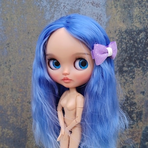 Puede incluir: Una muñeca de moda de color nude con pelo largo azul y ojos grandes azules. La muñeca tiene un pequeño lazo morado en el pelo. La muñeca está sujeta por un paño blanco. La cara de la muñeca tiene un rubor rosado suave.
