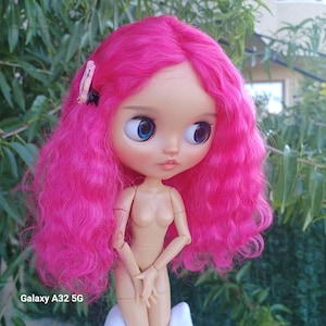May include: A doll with vibrant, wavy hot pink hair and large, round blue eyes. The doll has a pale complexion, a small nose, and pink lips. A small, rectangular hair clip with a colorful pattern is attached to the side of the hair.