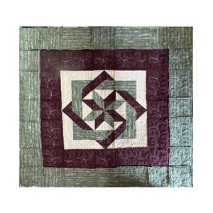 May include: A handmade quilt with a geometric design. The centre features a starburst pattern in shades of green, cream, and burgundy. The quilt is framed by a burgundy border with white floral stitching and a green outer border. The overall aesthetic is traditional.