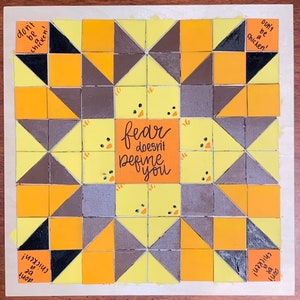 May include: A yellow, brown, and black quilt block with a geometric pattern. The center of the block features the text "fear doesn't define you". The block is surrounded by the text "don't be a chicken!" repeated four times.