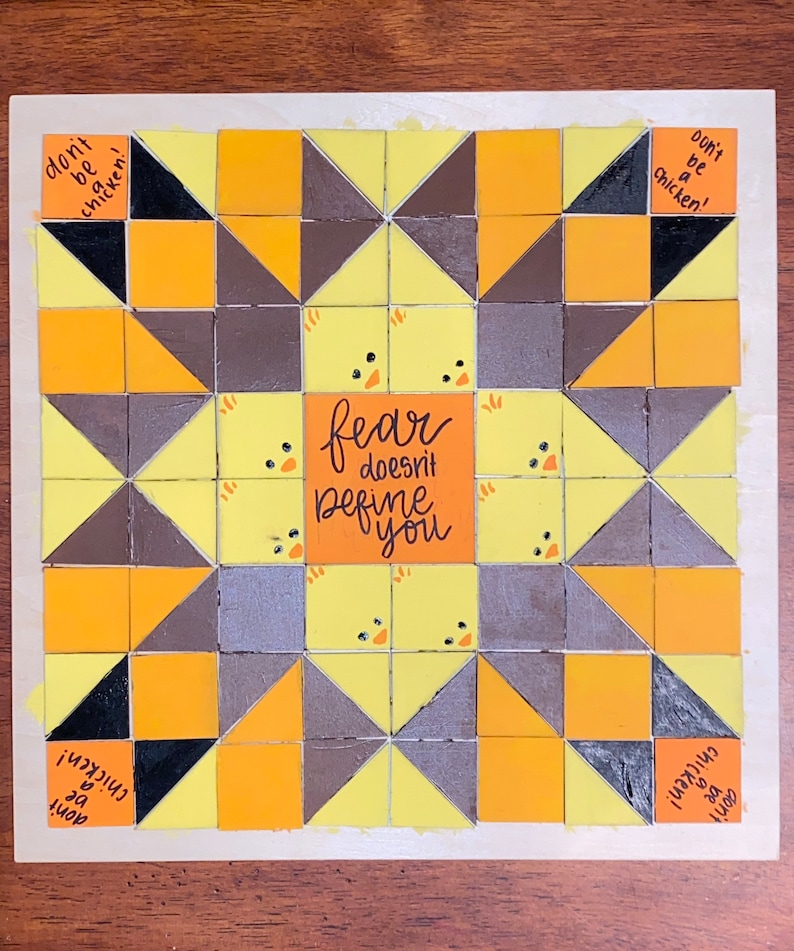 May include: A yellow, brown, and black quilt block with a geometric pattern. The center of the block features the text "fear doesn't define you". The block is surrounded by the text "don't be a chicken!" repeated four times.