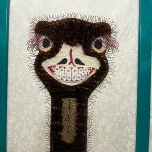 May include: Embroidered image of an ostrich with brown feathers and a long neck. The ostrich has a wide smile with large black eyes and a set of white teeth with green and red braces.