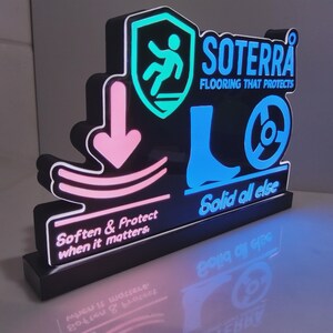 Può includere: Un'insegna LED nera con il logo SOTERRA e il testo Flooring That Protects. Mostra un'icona a scudo turchese con una persona che cade, una freccia rosa che punta a una linea curva e un'icona blu di un piede e una ruota con il testo Solid all else.