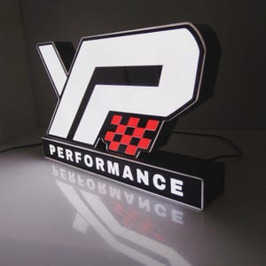 May include: A custom illuminated sign featuring the letters YP in white with a black border, accompanied by a red and black checkered square graphic. Below the letters, a rectangular base displays the word PERFORMANCE in white text.
