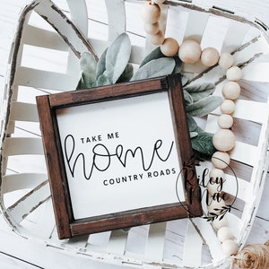 K&ouml;nnte beinhalten: Ein kleines, quadratisches Holzschild mit dem Text "TAKE ME HOME COUNTRY ROADS" in schwarzer Schrift. Das Schild ist in dunklem Braunholz gerahmt und liegt auf einem wei&szlig;en, gewebten Tablett mit dekorativem Gr&uuml;n und Holzperlen.