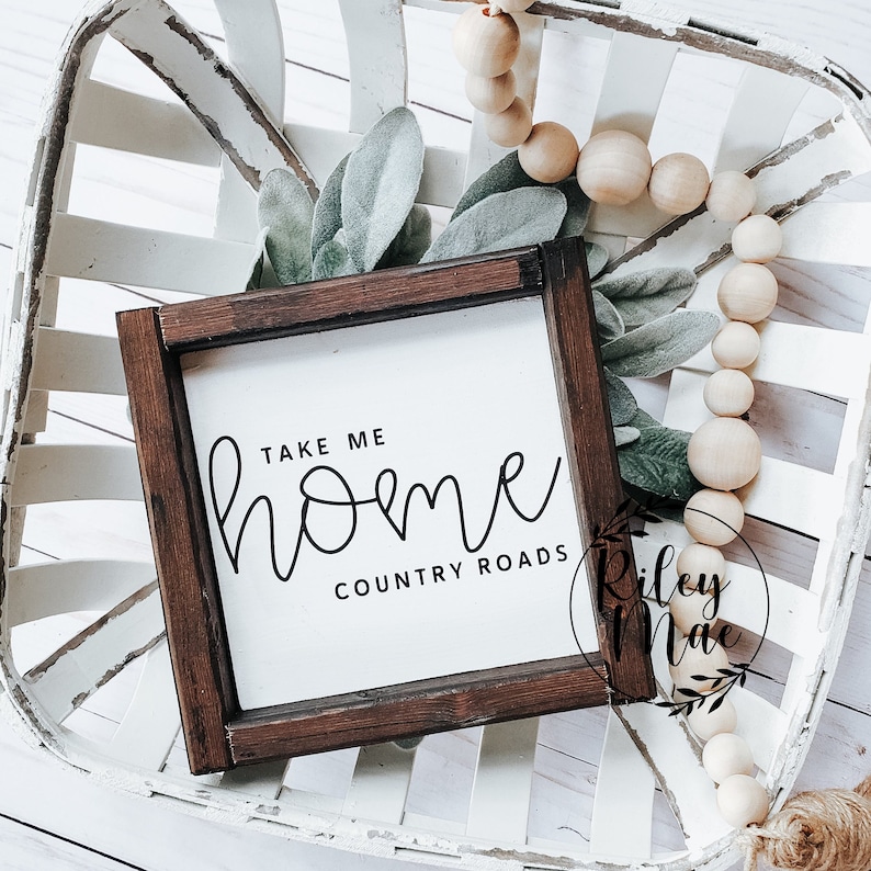 K&ouml;nnte beinhalten: Ein kleines, quadratisches Holzschild mit dem Text "TAKE ME HOME COUNTRY ROADS" in schwarzer Schrift. Das Schild ist in dunklem Braunholz gerahmt und liegt auf einem wei&szlig;en, gewebten Tablett mit dekorativem Gr&uuml;n und Holzperlen.