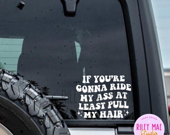 Może przedstawiać: Biała naklejka winylowa z napisem "IF YOU'RE GONNA RIDE MY ASS AT LEAST PULL MY HAIR" w stylu retro. Naklejka znajduje się na szybie pojazdu, z oponą widoczną na pierwszym planie. Naklejka zawiera również tekst "RILEY MAE STUDIO TRENDY TEES".