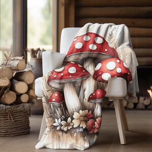 May include: A white throw blanket with a colorful illustration of red and white mushrooms with white spots, surrounded by brown and yellow flowers. The blanket is draped over a white chair with wooden legs.
