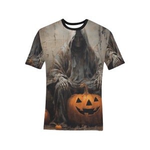 May include: A short-sleeved t-shirt featuring a Halloween-themed design. The graphic depicts a robed figure seated with a carved jack-o'-lantern. The color palette includes shades of gray, orange, and black, creating a spooky aesthetic.