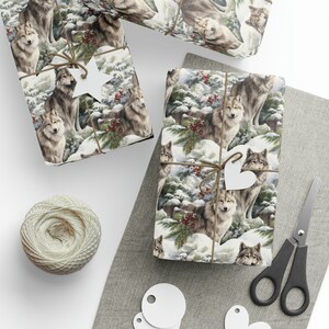 Op de afbeelding: Cadeaus verpakt met een winterwolfontwerp. Het cadeaupapier toont gedetailleerde illustraties van wolven in een besneeuwd bos met rode bessen. De cadeaus zijn vastgebonden met touw en versierd met witte hart- en stervormige labels. Een kluwen touw en een schaar zijn ook zichtbaar.