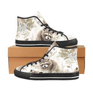 May include: High-top canvas sneakers featuring a raccoon illustration with floral accents in sepia tones. The shoes have black laces, eyelets, and a black rubber sole with a white stripe. The sneakers are displayed on a brown cardboard box.
