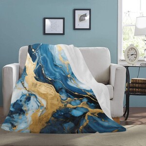May include: A soft, plush throw blanket with a blue and gold abstract marble pattern. The blanket is draped over a white couch in a living room setting.