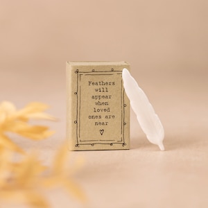 May include: A small, rectangular box with the text "Feathers will appear when loved ones are near" and a white feather. The box is brown with a decorative border. The feather is white and has a detailed design.