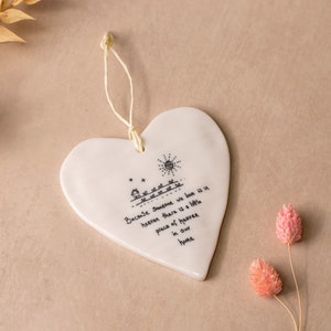 May include: A white heart-shaped ceramic ornament with a cream-colored string for hanging. The ornament features a black line drawing of a house, sun, and text that reads "Because someone we love is in heaven there is a little piece of heaven in our home."