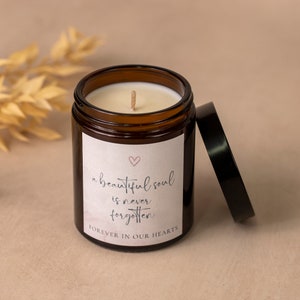 May include: A brown glass candle jar with a black lid, featuring a white label with the text "a beautiful soul is never forgotten" and a pink heart. The candle has a white wax and a wooden wick. The lid is placed next to the jar.