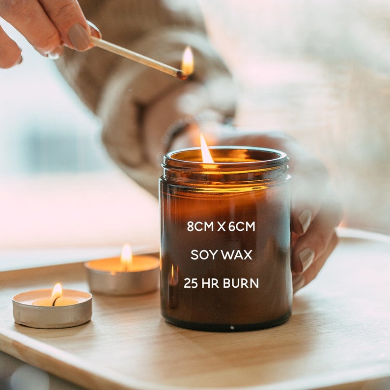 May include: A lit soy wax candle in a brown glass jar, measuring 8cm x 6cm, with a 25-hour burn time. A match is being used to light the candle. Two tea light candles are also lit on a wooden tray.