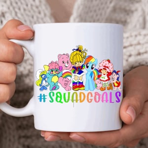 May include: White ceramic mug with a colorful illustration of cartoon characters including a rainbow unicorn, a Care Bear, a My Little Pony, and Strawberry Shortcake. The text "#squadgoals" is printed in rainbow colors below the characters.