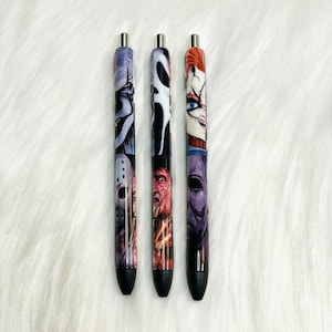 May include: Three black pens with different horror movie character designs. The pens feature Jason Voorhees, Ghostface, and Chucky.