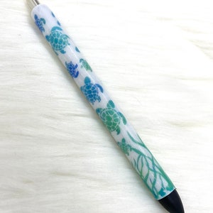 May include: A white pen with a blue and green sea turtle design. The pen has a black tip and a silver top.