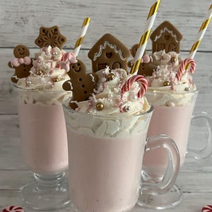 Puede incluir: Tres tazas de cristal llenas de una bebida rosa claro cubiertas con nata montada, galletas de jengibre con forma de casa y persona, pajitas a rayas doradas y blancas, bastones de caramelo pequeños y chispas doradas y plateadas.