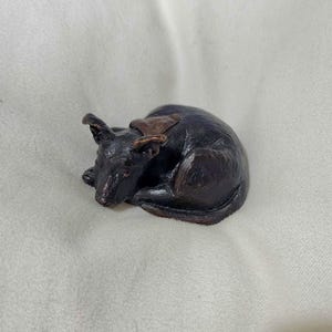 May include: A small bronze sculpture of a sleeping dog. The dog is curled up in a ball with its head resting on its paws. The sculpture is detailed and realistic.