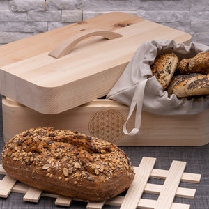 Puede incluir: Una caja de pan de madera con tapa abatible y asa. La caja tiene un diseño de flor en la parte delantera y una bolsa de lino en el interior con panecillos.