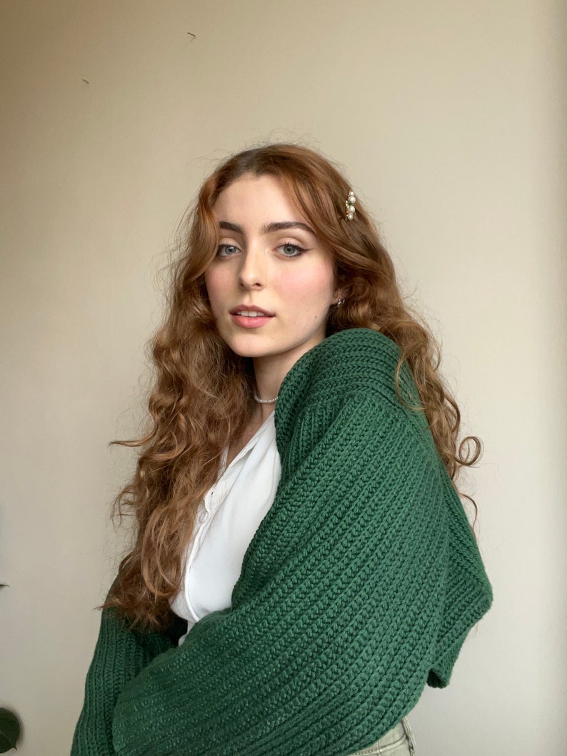 K&ouml;nnte beinhalten: Eine Frau mit langen, lockigen, rotbraunen Haaren tr&auml;gt einen dunkelgr&uuml;nen Strickcardigan. Sie tr&auml;gt ein wei&szlig;es Hemd unter dem Cardigan.