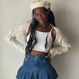 K&ouml;nnte beinhalten: Eine Person tr&auml;gt ein wei&szlig;es gestricktes Crop-Top mit einer wei&szlig;en Baskenm&uuml;tze und einen Jeansrock mit Falten.