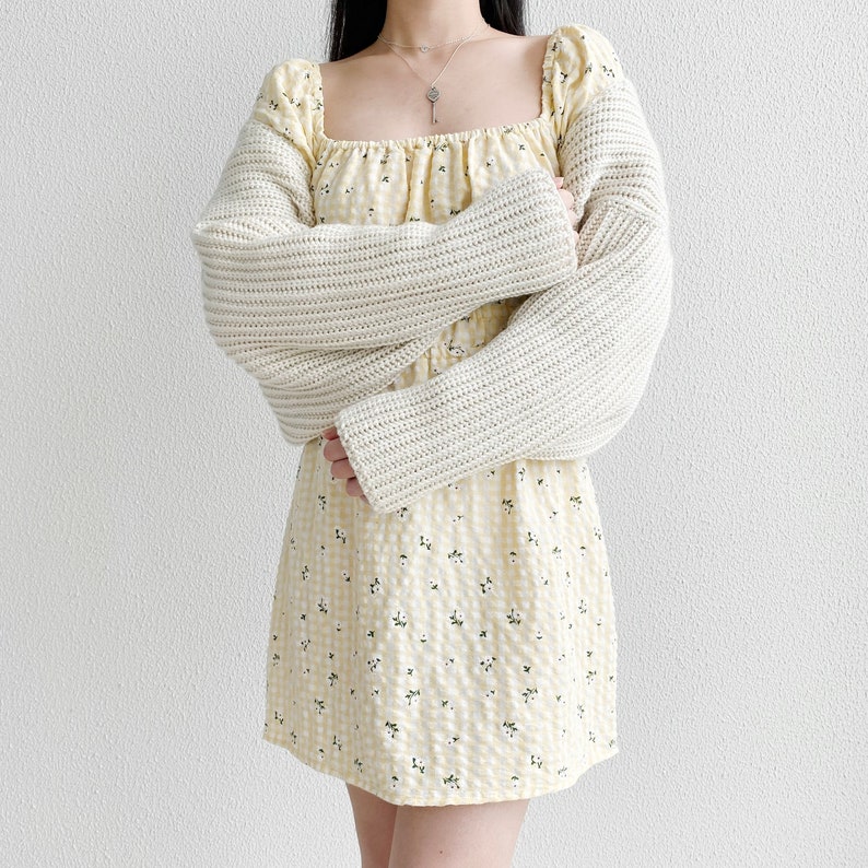 K&ouml;nnte beinhalten: Ein wei&szlig;er Strickcardigan mit langen &Auml;rmeln und quadratischem Ausschnitt, getragen &uuml;ber einem gelben Blumenkleid mit quadratischem Ausschnitt.