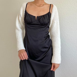 K&ouml;nnte beinhalten: Eine wei&szlig;e Strickjacke mit langen &Auml;rmeln und einem runden Ausschnitt. Die Jacke wird &uuml;ber einem schwarzen Satin-Slipdress getragen.