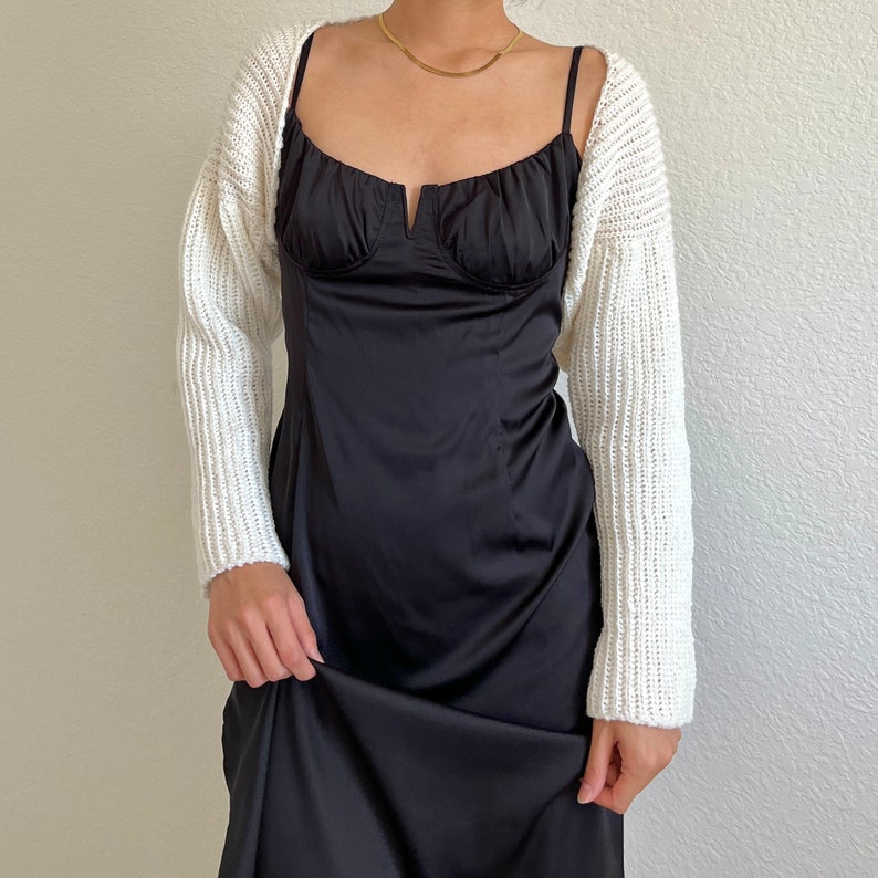 K&ouml;nnte beinhalten: Eine wei&szlig;e Strickjacke mit langen &Auml;rmeln und einem runden Ausschnitt. Die Jacke wird &uuml;ber einem schwarzen Satin-Slipdress getragen.