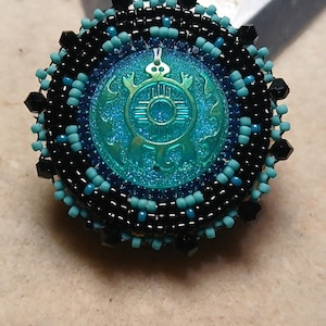 May include: A round, beaded pin with a turquoise and black color scheme. The center features a turquoise emblem with a turtle design. The pin is surrounded by black and turquoise beads, creating a detailed and decorative accessory.