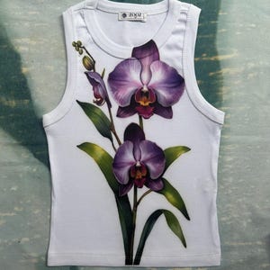 May include: A white ribbed vest top featuring a graphic print of two large purple orchids with green leaves and stems. The garment has a round neckline and a label at the inner collar that reads ZOOZ. The design is centred on the front of the shirt.