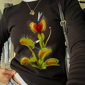 May include: Black long-sleeved top with a vibrant Venus flytrap illustration. The design incorporates green, red, and orange hues. The top is worn with blue denim jeans and a gold necklace.