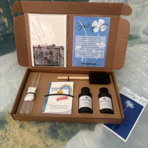 May include: An open cardboard box containing a cyanotype kit. The kit includes bottles of solution, pipettes, paper, stencils, and a guide. The box also contains a small booklet with a rainbow design and the text "Surprising with 2002".