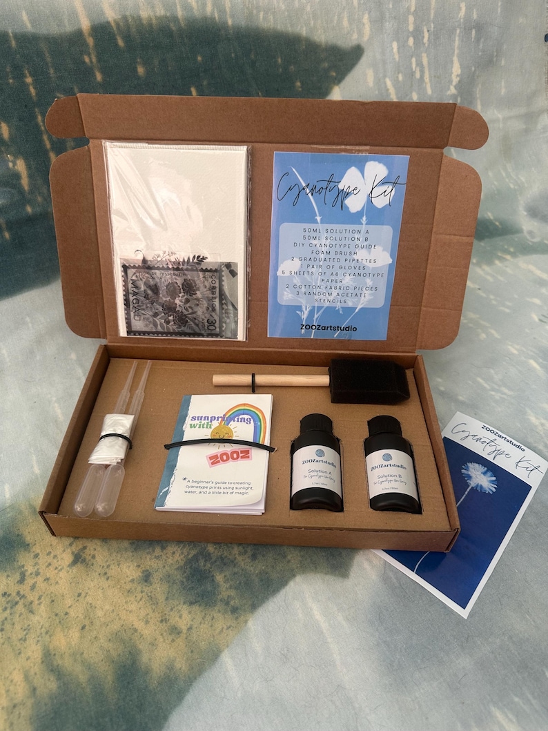 May include: An open cardboard box containing a cyanotype kit. The kit includes bottles of solution, pipettes, paper, stencils, and a guide. The box also contains a small booklet with a rainbow design and the text "Surprising with 2002".