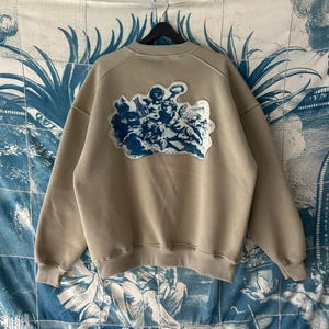 Pode incluir: Camisola bege com um desenho de querubins azul e branco nas costas. A camisola de gola redonda tem um corte folgado e mangas compridas. O desenho dos querubins é uma ilustração detalhada de várias figuras. A camisola está pendurada num cabide preto.