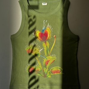 May include: Olive green ribbed tank top with a colorful Venus flytrap graphic. The graphic features a red heart-shaped center, yellow and green leaves, and a blue stem. The tank top has a small tag that says "zooz".