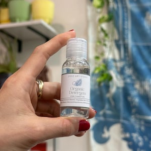 Puede incluir: Una pequeña botella transparente de detergente orgánico para cianotipia, sostenida en una mano. La etiqueta dice "Organic Detergent" y "ZOOZ ART STUDIO". La botella tiene una tapa blanca y una etiqueta blanca con un diseño de helecho.
