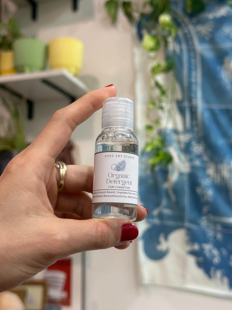 May include: A clear, small bottle of organic detergent for cyanotype, held in a hand. The label reads "Organic Detergent" and "ZOOZ ART STUDIO". The bottle has a white cap and a white label with a fern design.