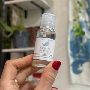 May include: A small, clear bottle of "Organic Detergent FOR CYANOTYPE" is held up. The label includes the text "ZOOZ ART STUDIO" and a botanical illustration. The bottle has a clear cap and contains a transparent liquid. The person holding the bottle has red nail polish.